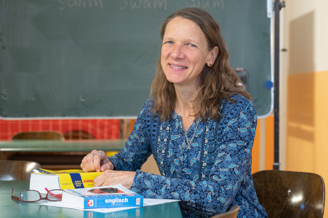 Eine Frau mit blauer Bluse und langen Haaren sitzt fröhlich lachend an einem Schreibtisch, vor ihr liegt ein Englischwörterbuch